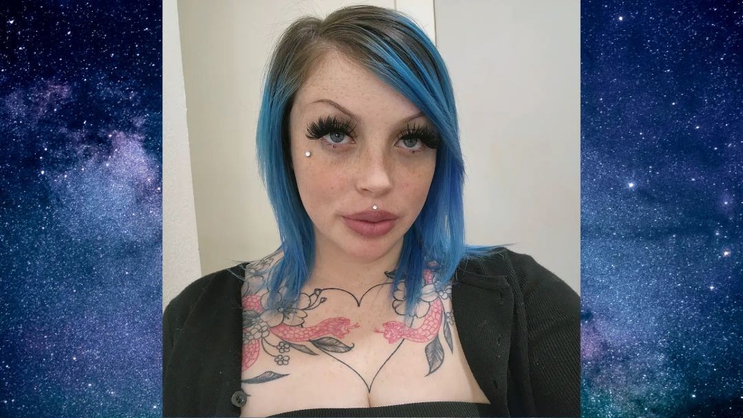 Person with blue hair, tattoos, and facial piercings against a starry background.