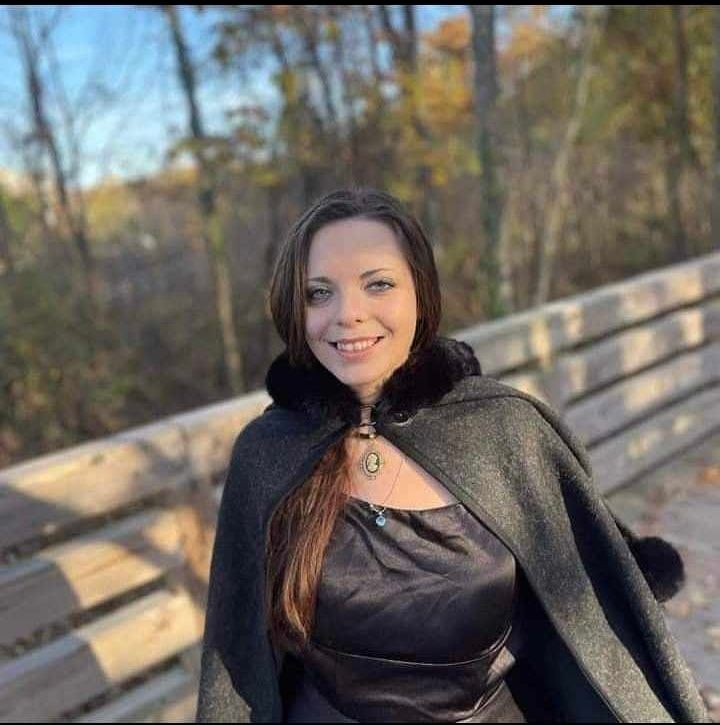 Woman in a dark cape smiling on a wooden bridge with a forest background.